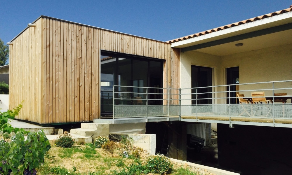 Extension moderne en bois avec bardage vertical, terrasse surélevée et large baie vitrée.
