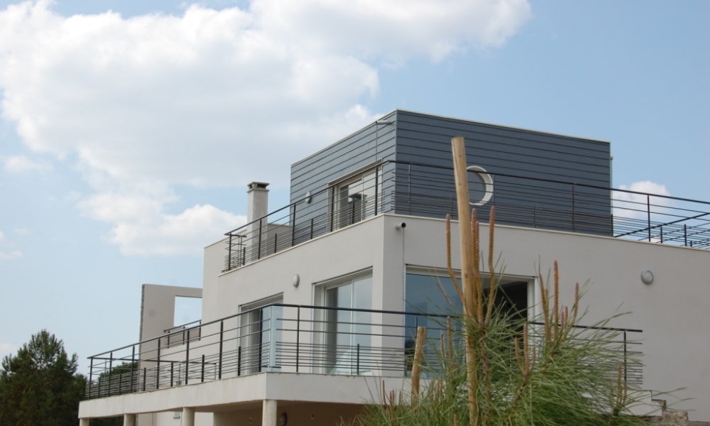 Maison moderne blanche avec toit-terrasse, garde-corps noirs et larges baies vitrées.