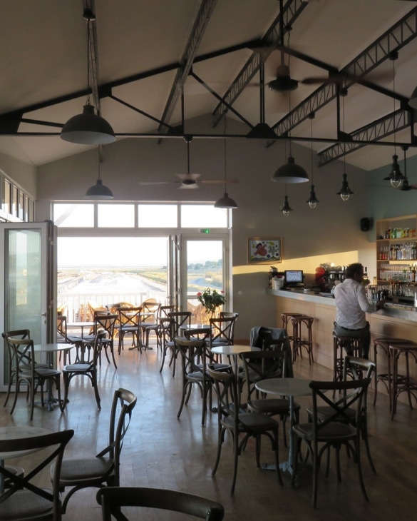 Intérieur d'un café lumineux avec mobilier bois, style industriel et terrasse ouverte sur un paysage naturel.
