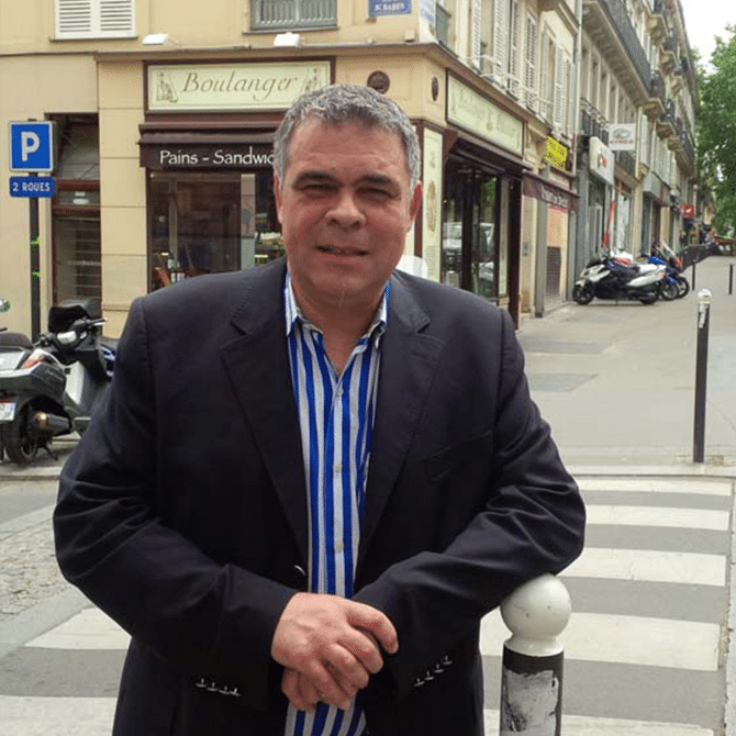 Homme en blazer souriant devant une boulangerie à Paris Homme souriant en blazer devant la boulangerie Pains-Sandwiches à Paris, près d'un passage piéton.