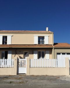 Maison beige, toit en tuiles, volets et clôture blancs. Numéro 379 sous un ciel bleu vif.