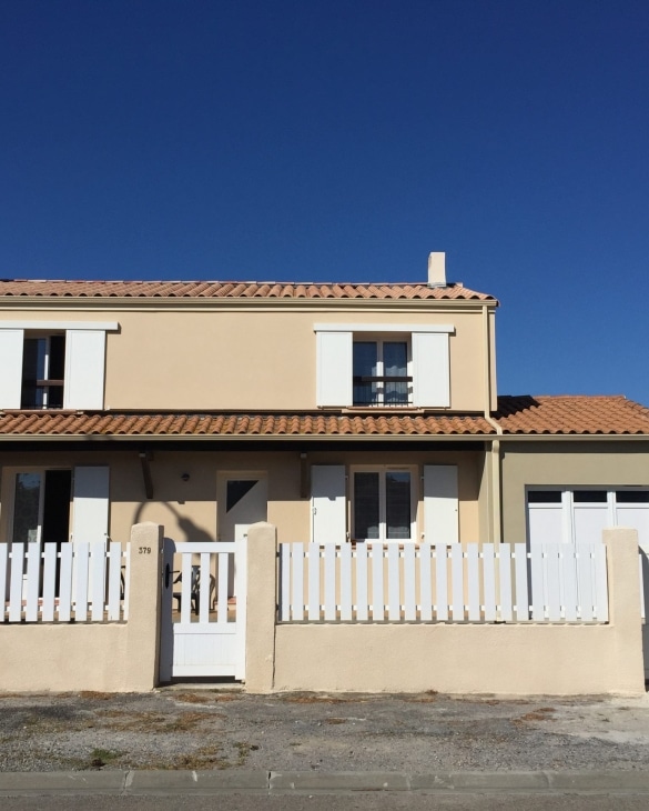 Maison beige, toit en tuiles, volets et clôture blancs. Numéro 379 sous un ciel bleu vif.