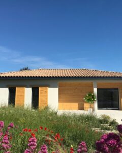 Maison à bardage bois et toit en tuiles nichée dans un jardin de fleurs sauvages sous un ciel bleu dégagé.