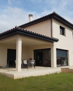 Maison moderne beige avec terrasse couverte, salon de jardin et toit en tuiles sombres sur pelouse.