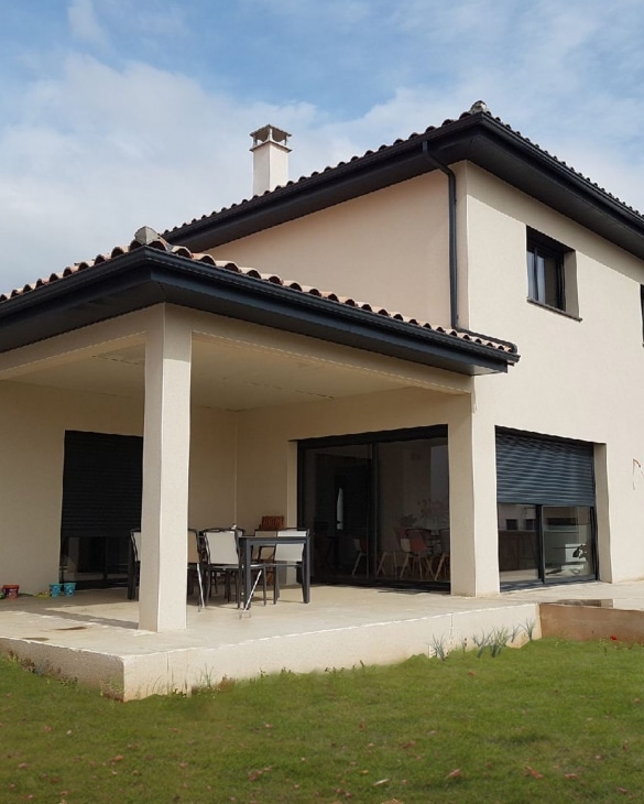 Maison moderne beige avec terrasse couverte, salon de jardin et toit en tuiles sombres sur pelouse.
