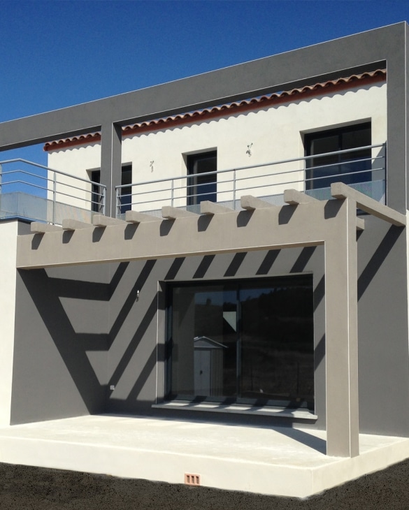 Façade maison moderne blanche et grise, pergola avec ombres géométriques sur terrasse ensoleillée.