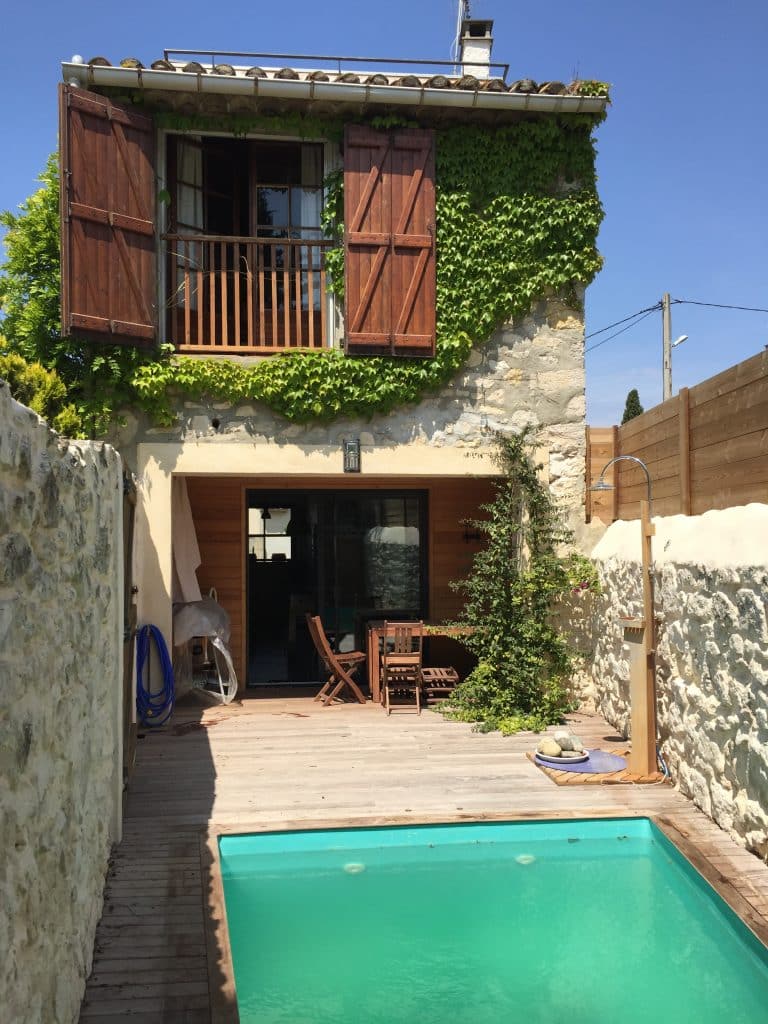 Maison en pierre, lierre, terrasse bois et petite piscine turquoise.