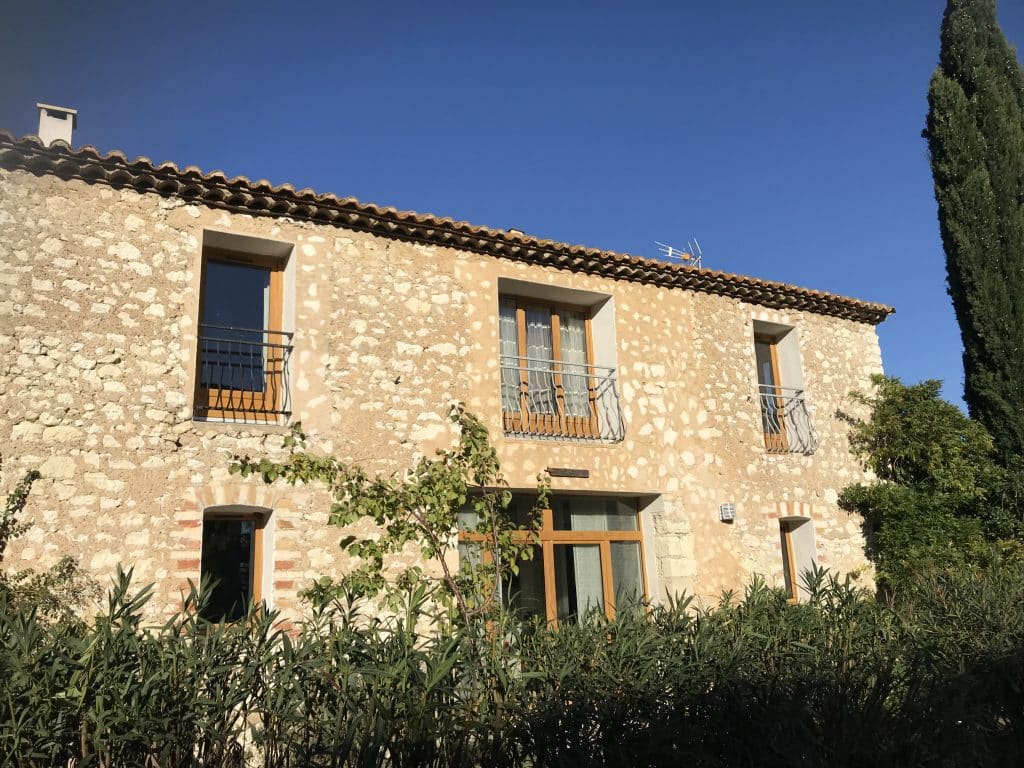 Maison en pierre provençale, fenêtres bois et fer forgé, végétation sous ciel bleu.