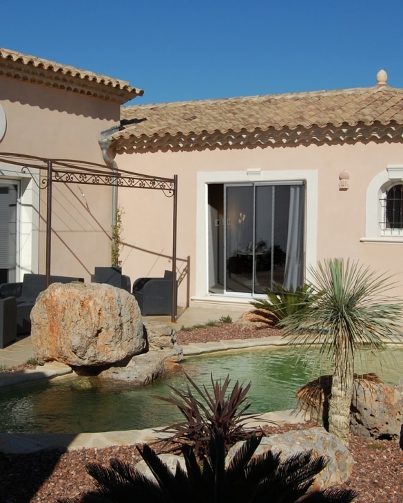 Maison méditerranéenne avec piscine, rochers, pergola en fer forgé et jardin exotique sous un ciel bleu.